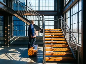 Jonge professional in marineblauw colbert beklimt moderne industriële trap van staal en hout, met geometrische architectuur en warm zonlicht