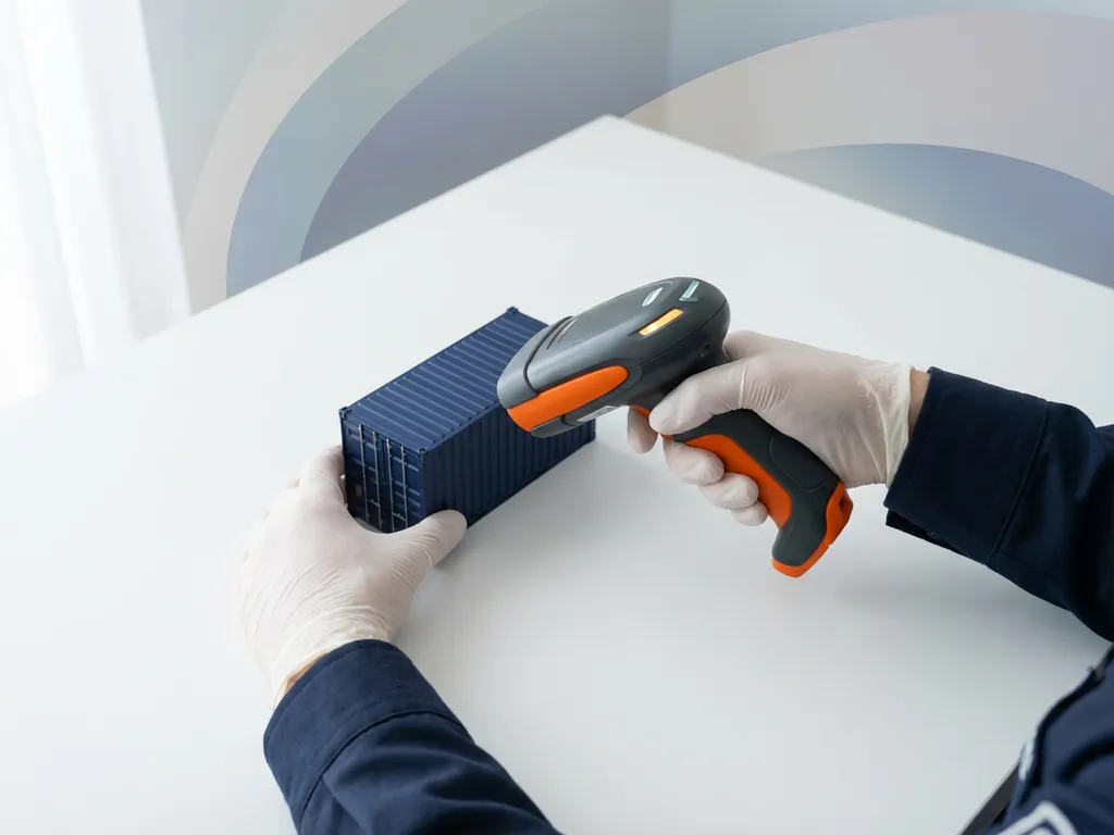 Handen van douanebeambte positioneren oranje handscanner boven donkerblauwe scheepscontainer op minimalistisch wit bureau.
