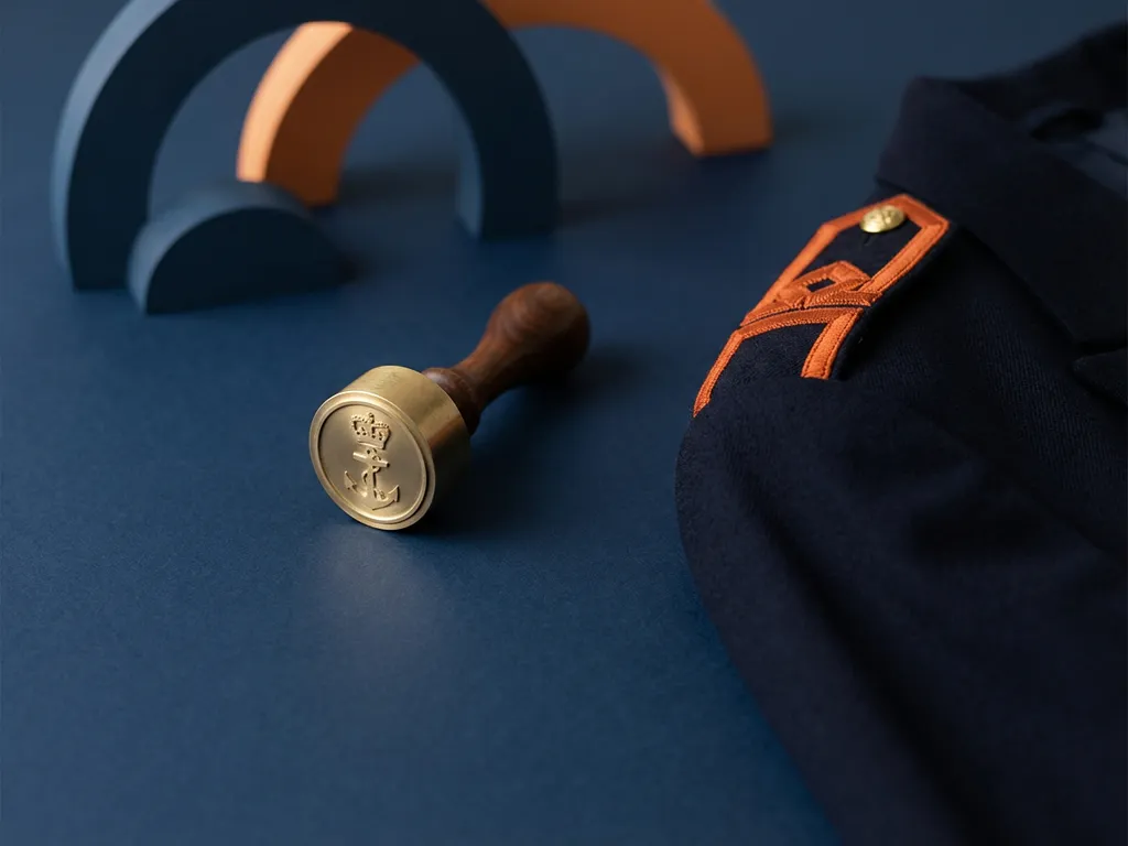 Koperen douanestempel naast gevouwen uniformmouw met oranje epaulet op marineblauw oppervlak, minimalistische flat lay compositie