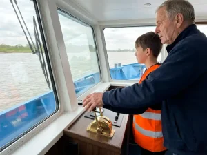 Ervaren kapitein begeleidt nieuwe bemanningslid bij messing stuurwielbediening in moderne binnenvaartschip stuurhut met uitzicht op rivier.
