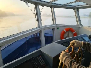 Verweerde handen van een kapitein aan een koperen stuurwiel in een moderne stuurhut, met uitzicht over een kalme rivier in ochtendlicht.