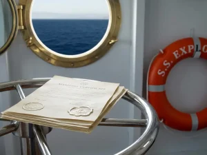 Stapel maritieme certificaten met reliëfzegels op een stalen stuurwiel, reddingsboei aan de wand en blauwe zee zichtbaar door patrijspoort.