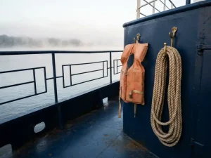 Verweerde oranje reddingsvest aan koperen haak naast opgerold meertouw op marineblauw scheepsdek, ochtendnevel boven kalm rivierwater.