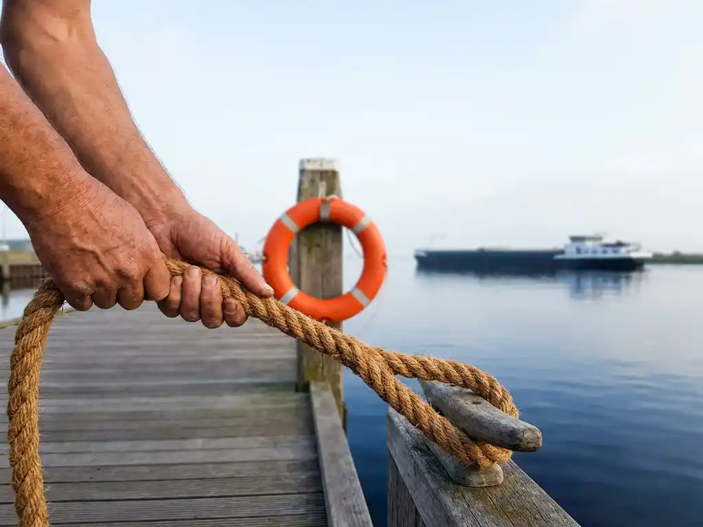 Verweerde handen grijpen een dikke meertros op een havenkade, oranje reddingsboei aan houten paal, vrachtschip op achtergrond