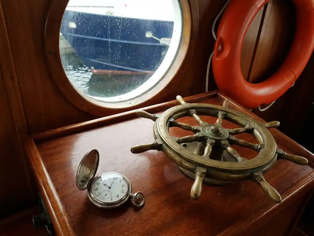 Verweerd koperen scheepsroer op mahoniehout stuurstand, antiek zakhorloge ernaast, reddingsboei aan kajuitwand, patrijspoort met waterdruppels.