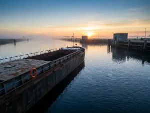 Verweerde vrachtschip glijdt bij dageraad over kalm kanaalwater, oranje reddingsboei aan de reling, gouden ochtendlicht werpt schaduwen