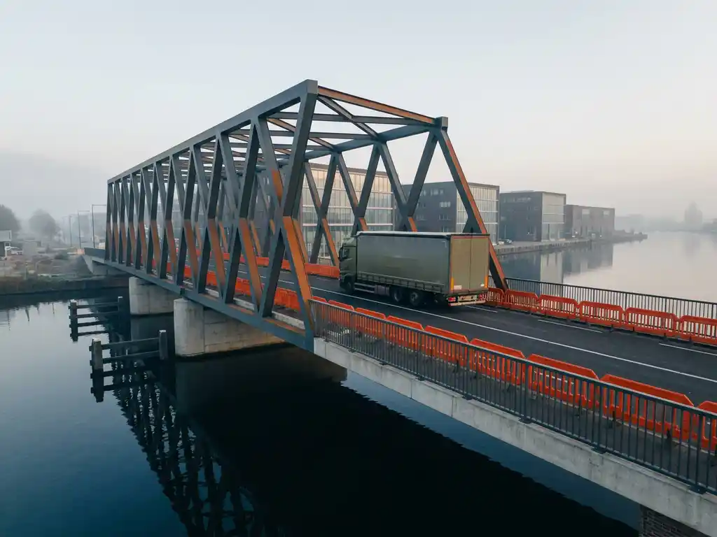 Vrachtwagen steekt moderne Amsterdamse grachtenbrug over richting pakhuiswijk, oranje veiligheidsbarrières langs donkerblauw water in zacht ochtendlicht.