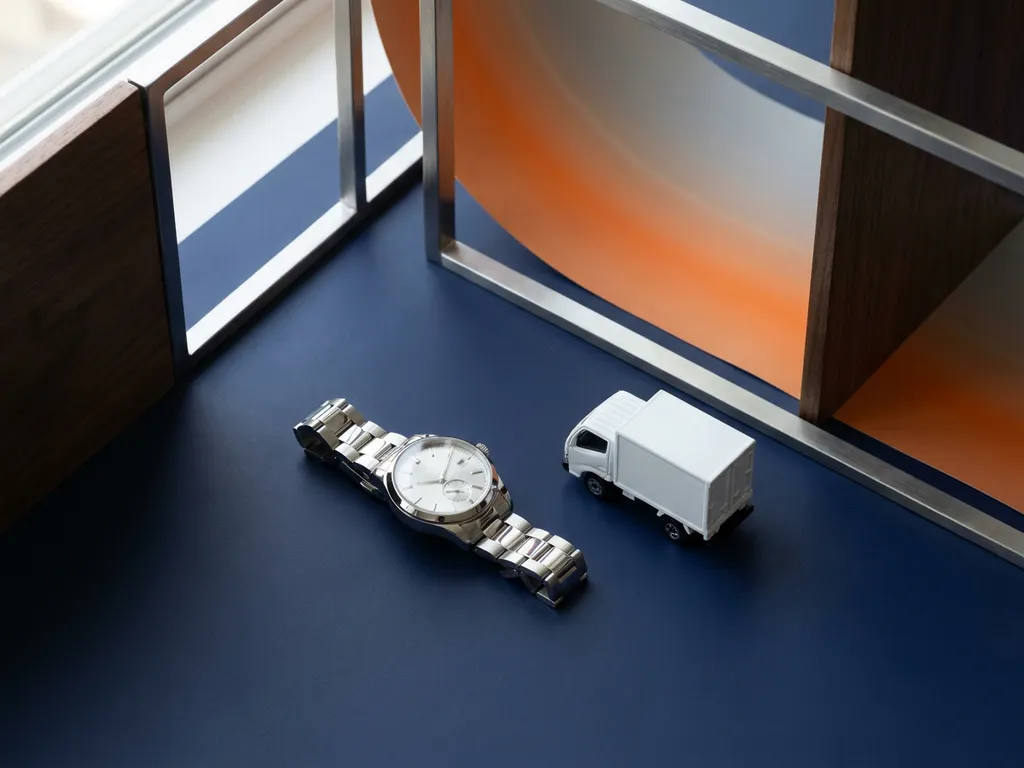 Chromen polshorloge naast miniatuur witte bestelwagen op donkerblauw oppervlak met oranje achtergrondaccent.