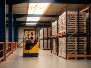 Heftruckchauffeur in oranje veiligheidsvest navigeert door magazijngang met blauwe stellingen en gestapelde pallets in natuurlijk licht.