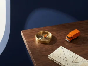 Koperen kompas met oranje naald naast miniatuur bezorgwagen en wegenkaart op donker houten bureau