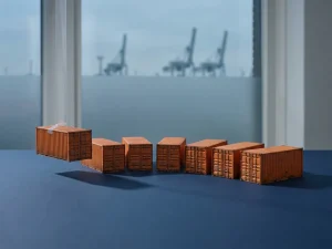 Miniatuur oranje zeecontainers in een boog gerangschikt op donkerblauw oppervlak, havenkranen van Rotterdam zichtbaar door matglas op de achtergrond.