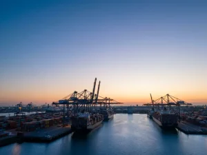 Containerkranen silhouetten tegen dageraadhemel bij Rotterdamse haven, gestapelde containers en vrachtschepen in kalm water.