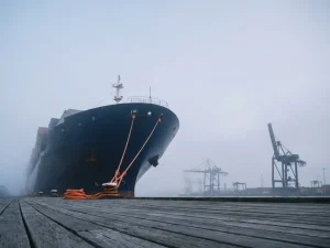 Boeg van marineblauw vrachtschip in ochtendnevel bij Rotterdamse haven, oranje meertouwen op verweerde kadeplanken, havenkranen op achtergrond.