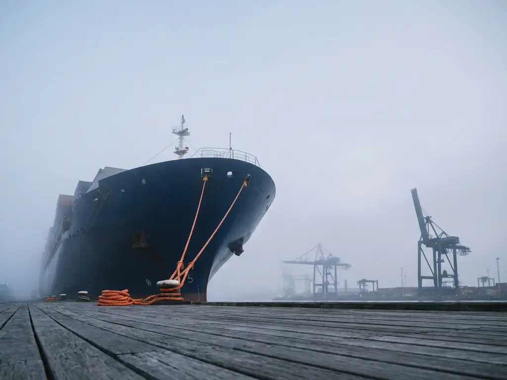 Boeg van marineblauw vrachtschip in ochtendnevel bij Rotterdamse haven, oranje meertouwen op verweerde kadeplanken, havenkranen op achtergrond.