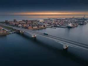 Vrachtauto rijdt over moderne brug tussen Nederlandse stad en internationale containerhaven bij zonsondergang, luchtfoto