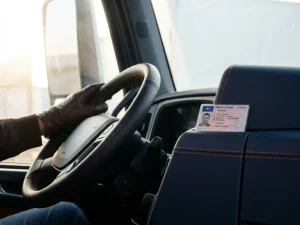 Gehandschoende hand op professioneel vrachtwagenstuur, rijbewijs op dashboard, marineblauw interieur met oranje stiksels in ochtendlicht.