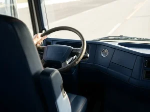 Vrachtwagenchauffeur houdt stuur vast in donkerblauwe cabine met oranje stiksels, analoge klok toont middag