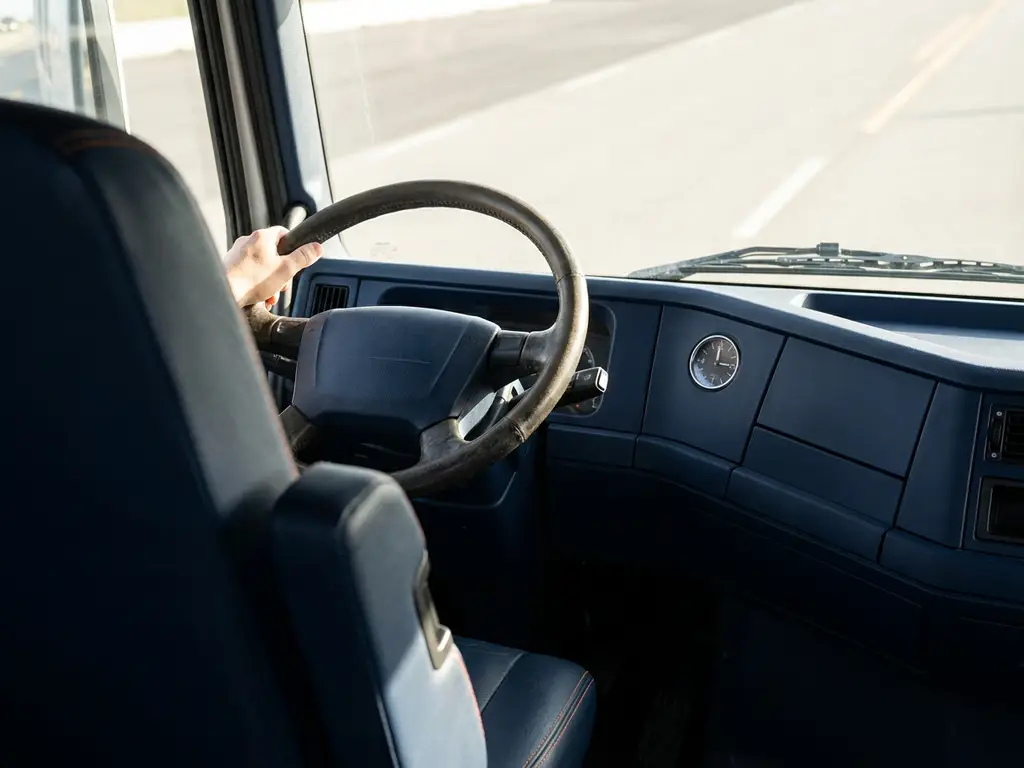 Vrachtwagenchauffeur houdt stuur vast in donkerblauwe cabine met oranje stiksels, analoge klok toont middag